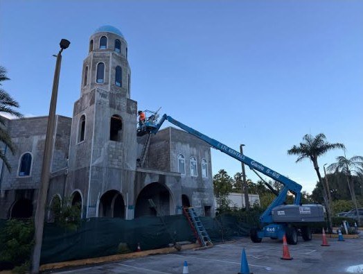 The Grand Masjid under construction: a worker in a raised boom lift beside the unfinished tower and its blue dome, with the arched arcade and the site fencing below