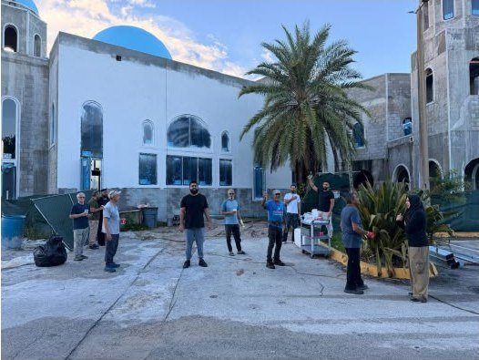 Volunteers standing in the courtyard between the finished white hall with its blue dome and the unfinished masjid wing, beside a palm tree, work carts and stacked building materials