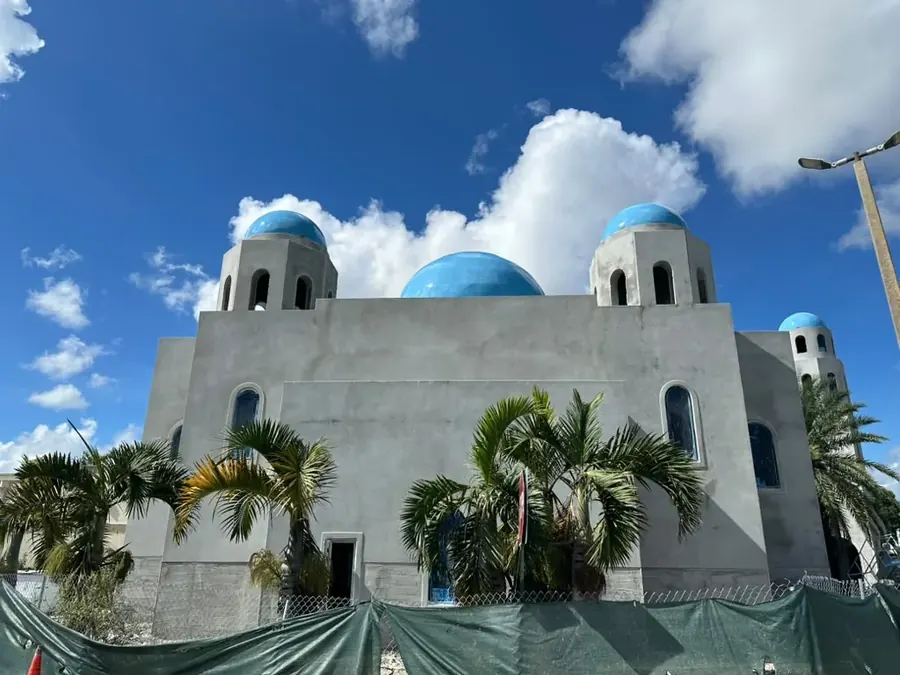 The Grand Masjid as it stands: the grey rendered exterior with four blue domes and arcaded corner towers, its arched windows glazed in blue tinted glass, behind palms and the site fencing
