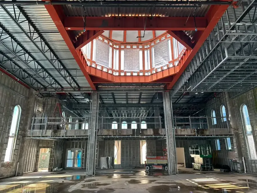 The main hall from the floor: the octagonal dome opening framed in orange steel above the ceiling joists, the mezzanine gallery and arched windows below, and a scissor lift standing on the bare concrete
